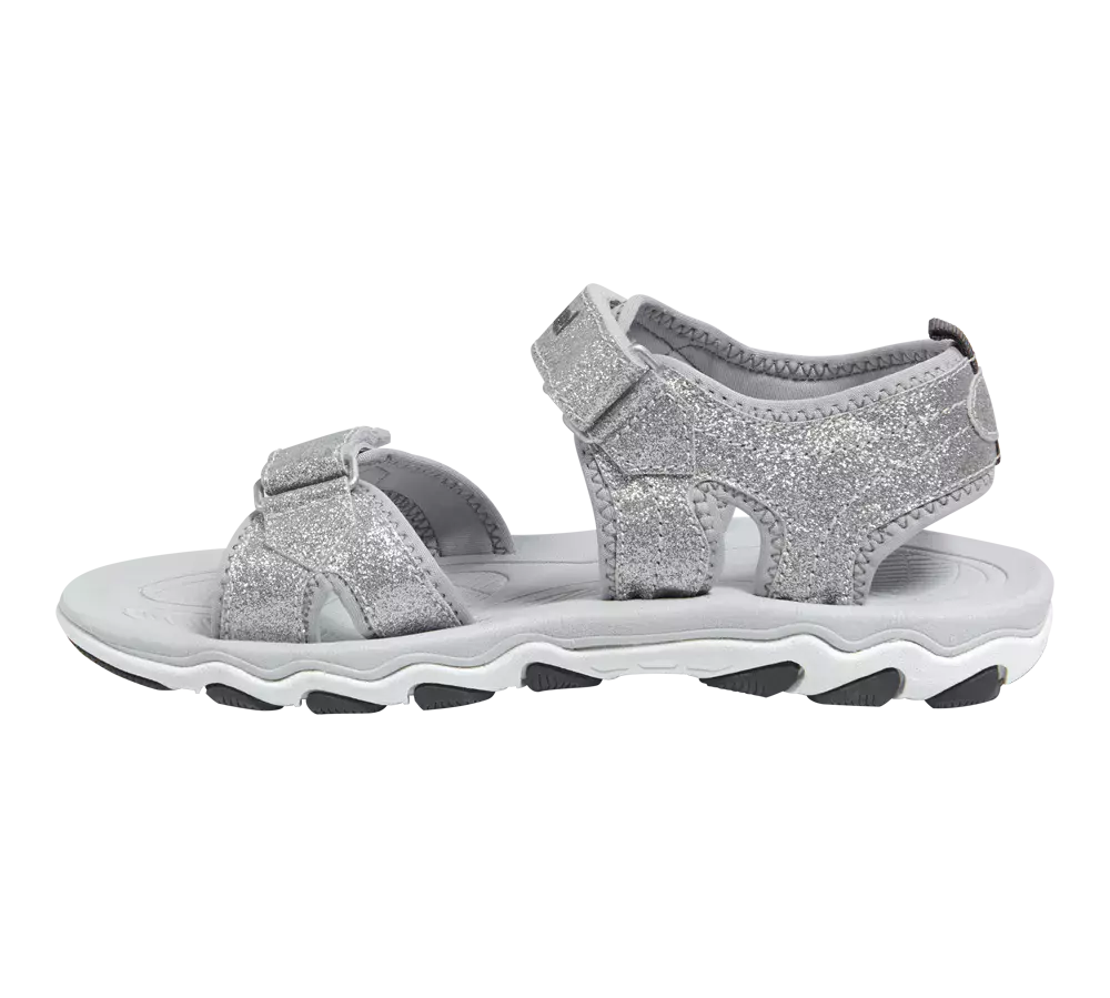 En sølvglitrende børnesandal med åbne tå- og hælkonstruktion, lavet med bløde, polstrede stropper og praktiske velcro-lukninger for justerbar pasform. Overfladen er dækket af fint glitter, mens indersålen virker polstret for komfort. Den robuste, bølgede gummisål i hvid og grå giver godt greb og stødabsorbering. Sandalen passer til både hverdag og festlige sommerlejligheder, hvor holdbarhed kombineres med et funklende look.