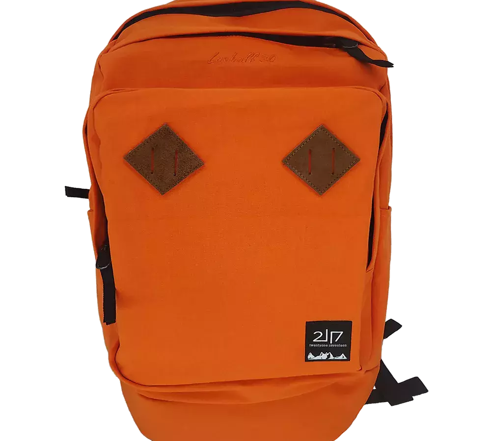 En klar orange rygsæk i robust lærredsstof med et enkelt, funktionelt design. Fronten har to brune diamantformede læderlapper, en lynlåslomme øverst og en rummelig hovedlomme. Sorte lynlåse og justerbare skulderstropper giver kontrast, mens et lille sort logoemblem nederst signalerer mærket. Rygsækken virker praktisk til skole, pendling eller dagsture med god opbevaringsplads og et tidløst, sporty udtryk.