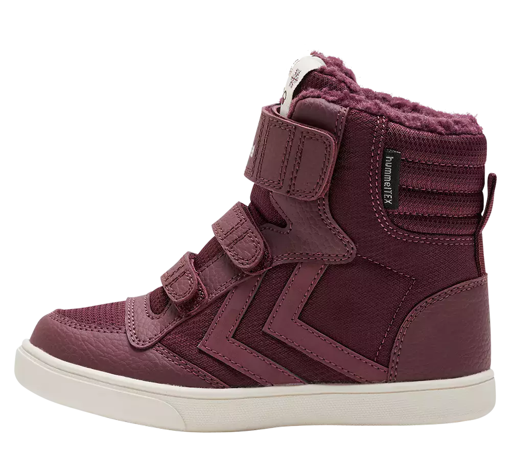 hindbærrødt højtop-sneakersstøvle til børn med blødt, pelsforet skaft. Overdelen kombinerer tekstureret læder og stofpaneler med dekorative syninger og et karakteristisk zigzag-motiv langs siden. Skoen lukkes med tre praktiske velcroremme, og en mærkeetiket "hummelTEX" sidder ved anklen. Den lyse, kraftige gummisål giver god støtte og kontrast, hvilket gør støvlen både varm, komfortabel og velegnet til hverdag.