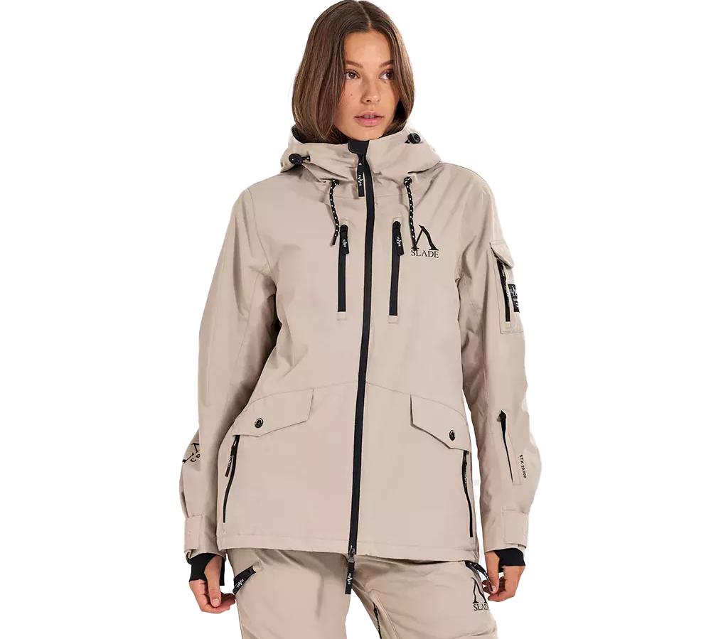 En model iført en lys beige, teknisk outdoorjakke med hætte og sorte kontrastdetaljer. Jakken har to vertikale lynlåse foran, flaplommer med knapper, håndledsmanchetter og justerbare snore i hætten. Matchende beige bukser giver et samlet look. Samlet fremstår outfittet funktionelt og stilrent, velegnet til udendørs aktiviteter med fokus på komfort og praktiske lommer. Detaljerne i sorte lynlåse og snore giver et moderne præg.