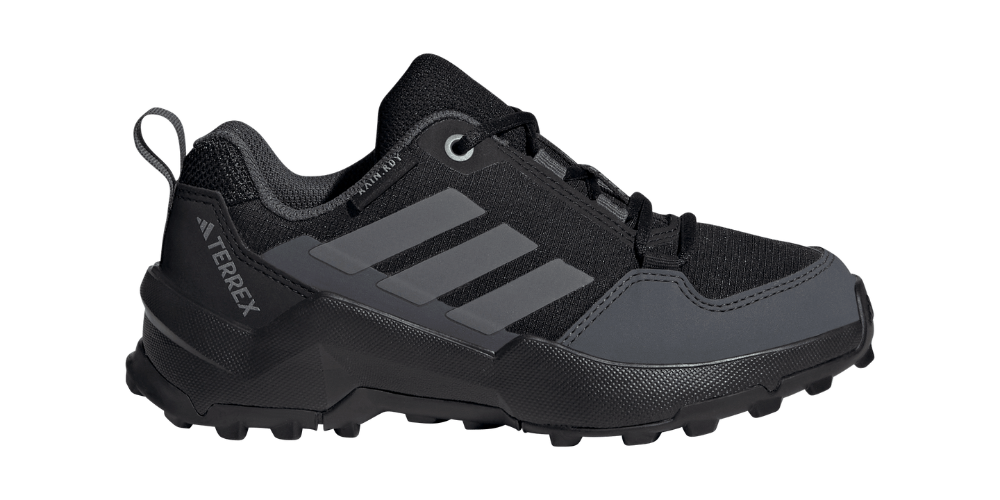 Et lavtstående, robust trail-løbesko i mørkegrå og sort med et funktionelt, minimalistisk design. Skoen har forstærkede paneler ved tå og hæl, åndbart mesh, polstret krave og klassiske tre striber langs siden. Aggressivt mønstret ydersål sikrer greb på ujævnt underlag, mens snørelukning og en bageste trækkestrop giver stabil pasform og praktisk påtagning. Branding 'Terrex' signalerer terrænorienteret design og holdbarhed.
