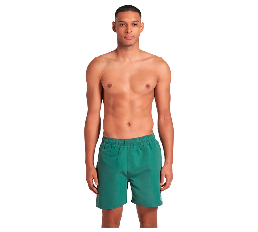 En mand står oprejst og kigger lige frem med et neutralt, roligt ansigtsudtryk. Han er skjorteløs og har en atletisk, slank overkrop med synlige muskler. Han bærer grønne bade- eller træningsshorts med elastisk talje, hænderne hviler afslappet langs siden. Baggrunden er neutral og ensfarvet, hvilket fremhæver motivets simple, minimalistiske og sportslige fremtoning.