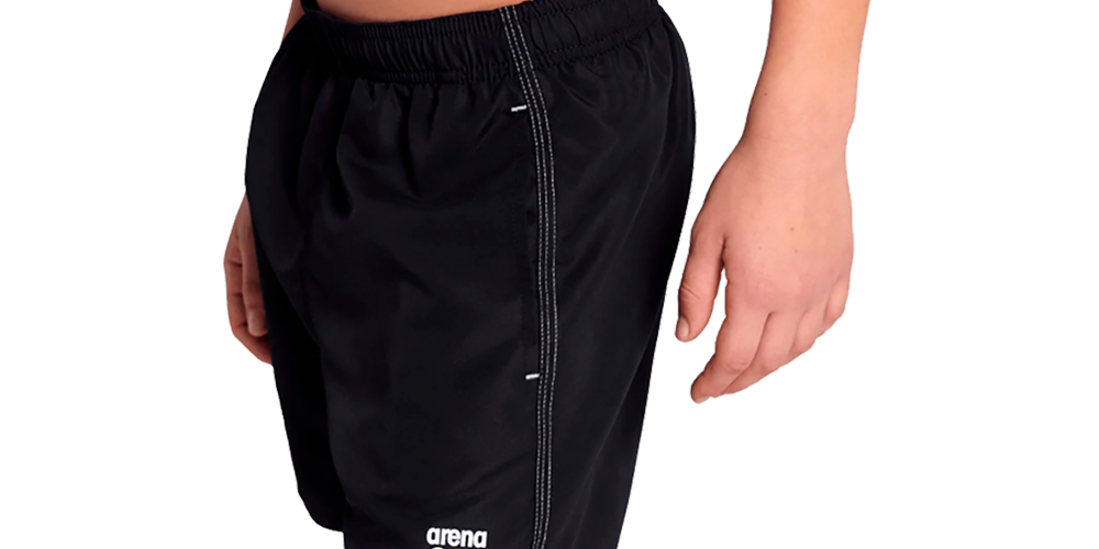 Billedet viser en ung person iført sorte badeshorts fra Arena. Shortsene har elastisk talje, knælængde pasform og et lille hvidt logo nederst på venstre ben. Siderne har synlige kontrastsømme og en enkel, sporty silhuet. Materialet virker let og hurtigtørrende, velegnet til svømning eller strandbrug. Farven er klassisk sort, hvilket giver et tidløst og neutralt udtryk.