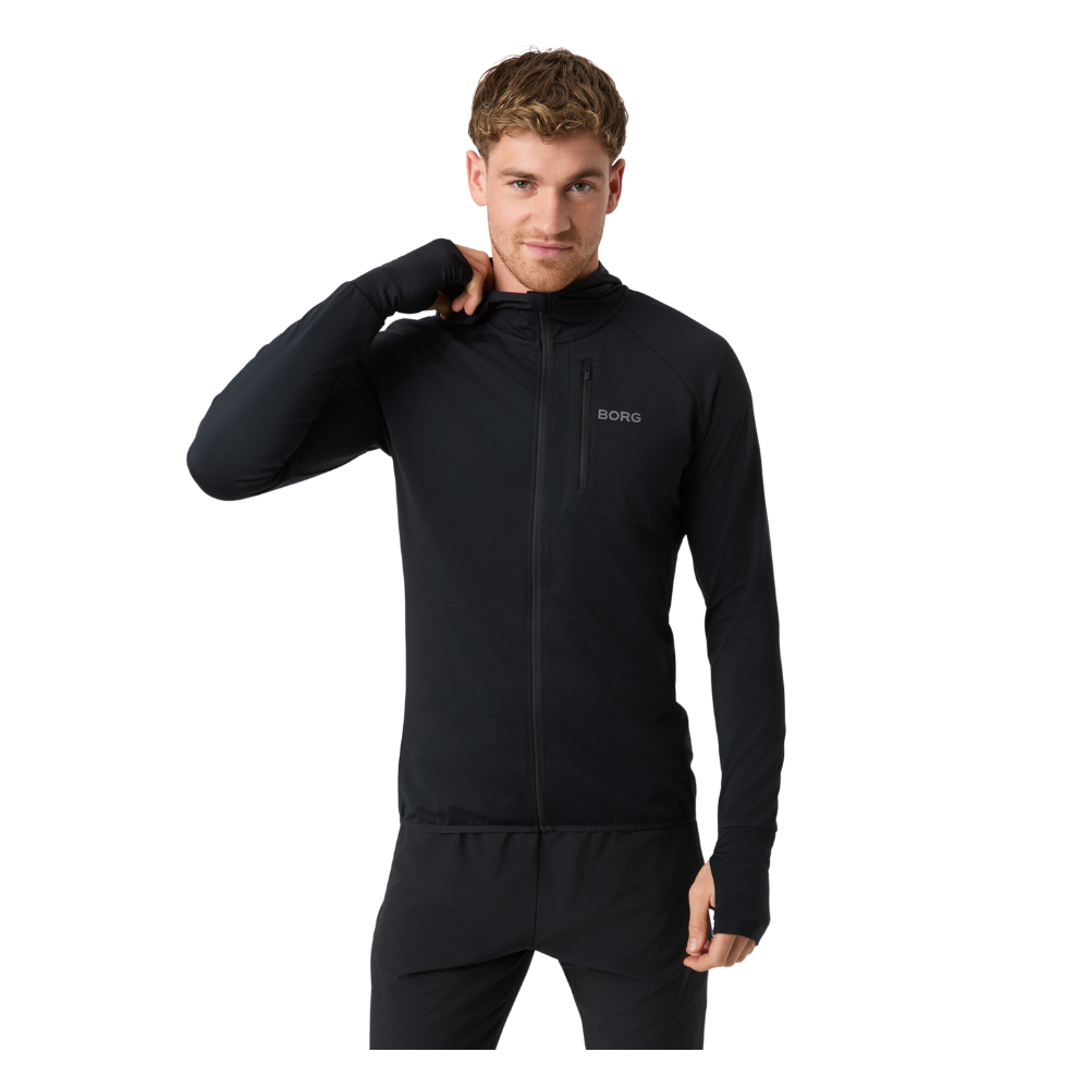 En ung mand iført en sort, tætsiddende træningsjakke med fuld lynlås og brystlomme med lynlås. Han står afslappet med den ene hånd ved nakken og et mildt smil, klar til løb eller træning. Jakken har lange ærmer med tommelfingerhul og et minimalistisk logo på brystet. Helhedsindtrykket er moderne, funktionelt og sporty.