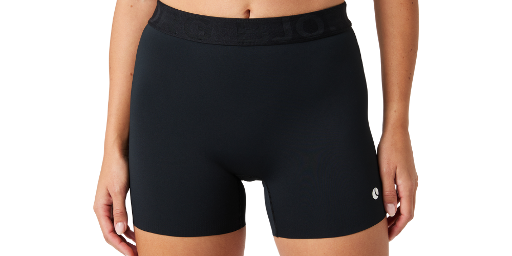 Billedet viser en person i en nærbilledet fremstilling af mave og lår iført sorte træningsshorts og en sort sports-bh. Shortsen har en bred elastik i taljen med diskret logo og et lille hvidt ikon på benet. Motivets hud er solbrun og velplejet, poseringen er afslappet med hænderne langs siden. Stilen fremstår enkel, sporty og funktionel med et rent, studiopræget udtryk.