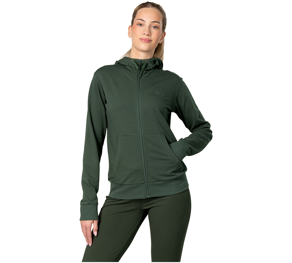 Billedet viser en kvinde iført en mørkegrøn, lynlåstrøje med hætte og matchende leggings. Hun står afslappet med den ene hånd i lommen og et let smil, mens håret er trukket tilbage. Tøjet har rene linjer, ribkanter og en praktisk kængurulomme, hvilket giver et sporty og komfortabelt udtryk egnet til træning eller hverdag.