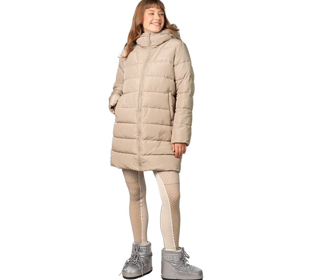 En kvinde iført en lang, beige dynejakke med hætte står afslappet og smilende. Jakken er tætsyet i vandrette baner og når til knæene. Hun har mønstrede, lyse strømpebukser og sølvglinsende Moon Boot-støvler, der giver et vinterligt udtryk. Hendes hænder er i lommerne, og helhedsindtrykket er varmt, moderne og funktionelt til kolde dage.