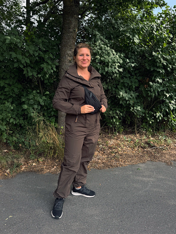 Kvinde i brun jakke og bukser med hætte, sort tværgående taske og sorte sneakers på asfalt foran træ.