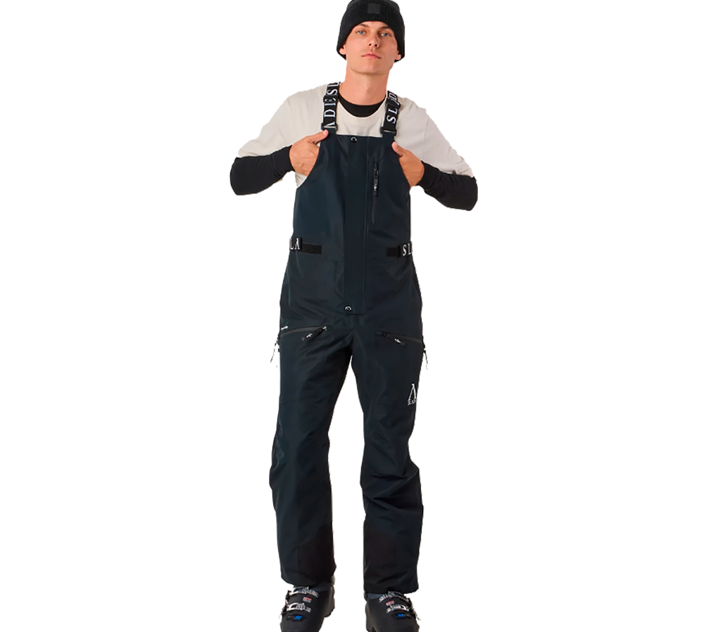 På billedet ses en ung mand iført Slade Aspen Bib Pants, som er designet til skisport. Disse mørkeblå skibukser fremstår med justerbare seler, der tydeligvis sikrer en behagelig pasform. Bukserne er udstyret med flere lynlåslommer, praktisk til opbevaring under aktivitet. Samlet set kombinerer bukserne funktionalitet med et moderne snit, perfekt til den stilbevidste skiløber.