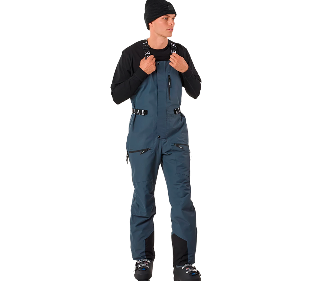 På billedet ses en ung mand, der præsenterer Slade Aspen Bib Pants, som er designet til mænd. Buksen har justerbare seler, flere funktionelle lommer, og den blå farve fremhæver et moderne snit. Skibukserne fra Slade kombinerer stil med praktiske detaljer, hvilket gør dem ideelle til skisport og vinteraktiviteter. Detaljerne og kvaliteten sikrer holdbarhed og komfort i koldt vejr.