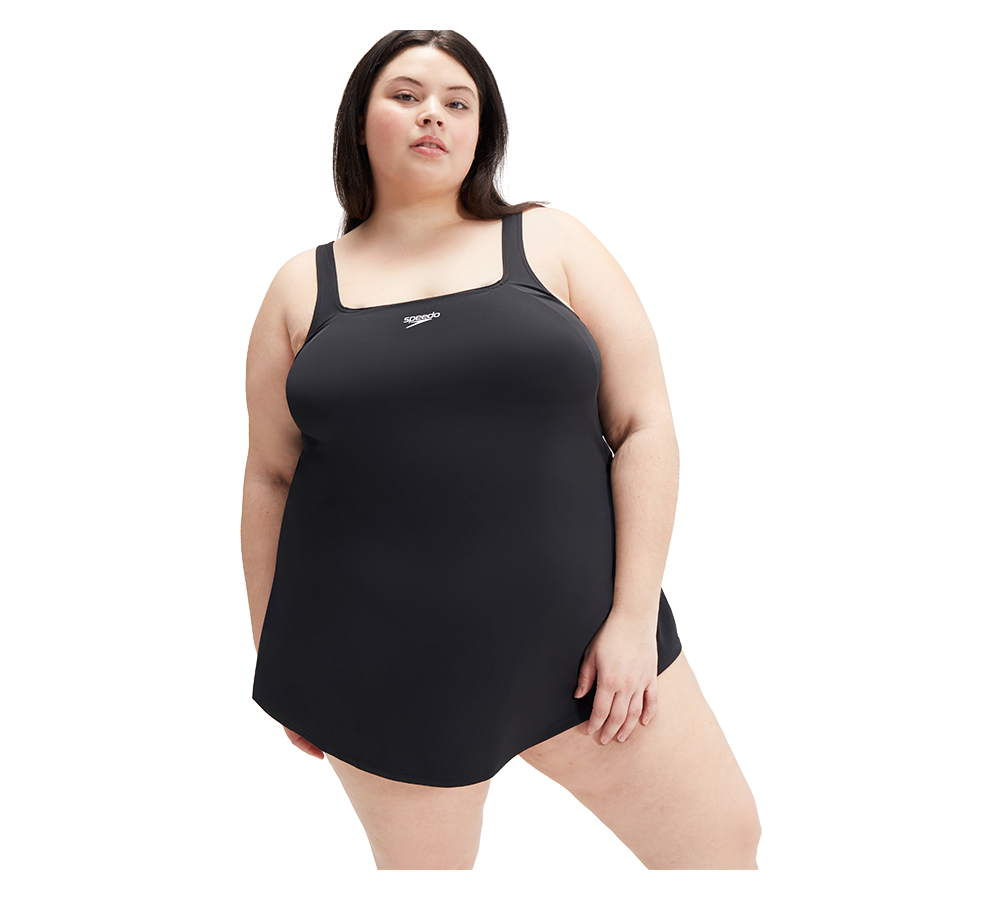 På billedet ses en person iført en enkel, sort badedragt med skørt (swimdress) og brede stropper. Modellen står i en selvsikker, afslappet positur mod en neutral, hvid baggrund. Badedragten har firkantet halsudskæring og et lille logo foran, hvilket giver et klassisk og funktionelt udtryk. Billedet udstråler komfort, inkludering og en tidløs stil, velegnet til både svømning og afslapning ved vandet.