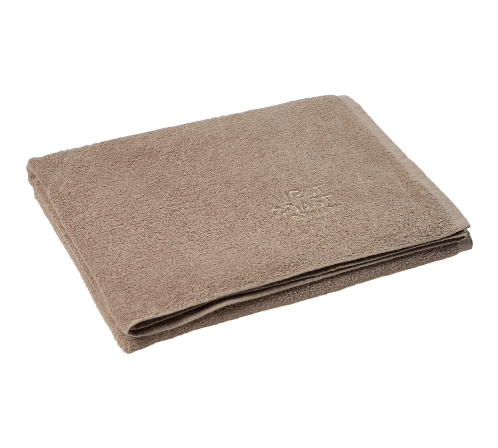 Et blødt, beige badehåndklæde er omhyggeligt foldet og lagt fladt mod en lys baggrund. Stoffet fremstår fyldigt og velplejet med tætte bomuldstresser, og en diskret indvævet tekstur og kantafslutning giver et enkelt, elegant udtryk. Håndklædet virker absorberende og komfortabelt, velegnet til både badeværelse og spa, og signalerer høj kvalitet og roligt design.