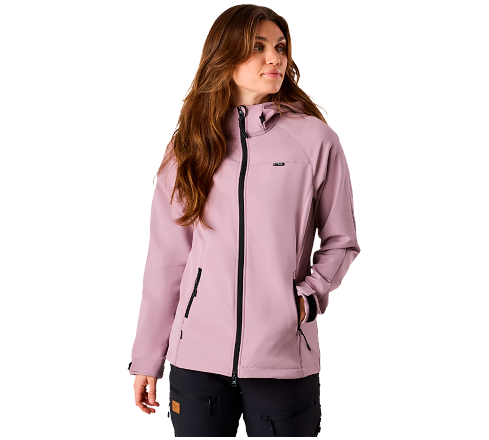 En kvinde iført en lyserosa softshell-jakke står i afslappet positur med hænderne i lommerne, vendt let mod højre. Jakken har sort lynlås og praktiske sidelommer, og sidder tæt ved hoften. Hendes langt, brunt hår falder løst over skuldrene, og hun giver udtryk for rolig opmærksomhed. Billedet fremhæver jakken som moderne, funktionel og velegnet til udendørs brug.
