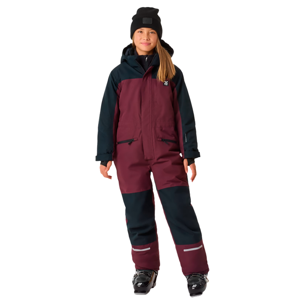 På billedet ses en ung pige iført X-TRAIL Cortina Overall Jr, en praktisk børne flyverdragt fra X-Trail. Dragten er tonet i mørke nuancer af rød og blå, hvilket giver et stilrent og holdbart look. Den er designet til at holde børn varme og tørre i alt slags vejr, perfekt til udendørs aktiviteter i vinterperioden. Dragtens robuste materiale og komfortable pasform sikrer, at børn kan bevæge sig frit og ubesværet uanset vejret.