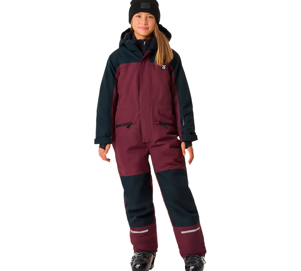 På billedet ses en ung pige iført X-TRAIL Cortina Overall Jr, en praktisk børne flyverdragt fra X-Trail. Dragten er tonet i mørke nuancer af rød og blå, hvilket giver et stilrent og holdbart look. Den er designet til at holde børn varme og tørre i alt slags vejr, perfekt til udendørs aktiviteter i vinterperioden. Dragtens robuste materiale og komfortable pasform sikrer, at børn kan bevæge sig frit og ubesværet uanset vejret.