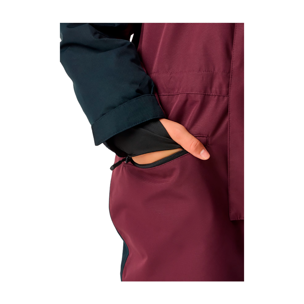 Billedet viser en ung pige iført X-TRAIL Cortina Overall Jr., en børneflyverdragt fra mærket X-Trail. Dragten primært i mørkerødt med detaljer i mørkeblå indeholder justerbare stropper og forstærkninger ved benene, perfekt for vinteraktiviteter. Den kombinerer funktionalitet med stil og sikrer varme og komfort i kølige vejrforhold. Modellen ser glad og tilpas ud i det udendørs outfit, der lover holdbarhed og bevægelsesfrihed.