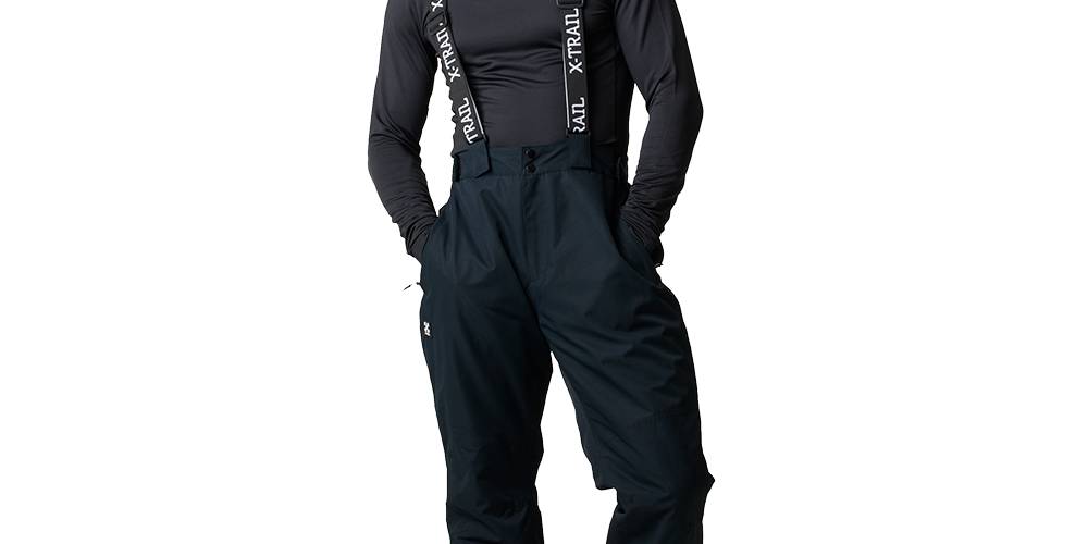En mand i afslappet positur står iført sort langærmet trøje og mørke, vatterede skibukser med seler, hvor teksten X-TRAIL ses tydeligt. Han har krøllet, mørkt hår og kigger direkte mod kameraet med en neutral mine. Bukserne er rummelige med sidelommer, og hans hænder er i lommerne. Robuste støvler fuldender det funktionelle, sporty outfit til udendørs aktiviteter.