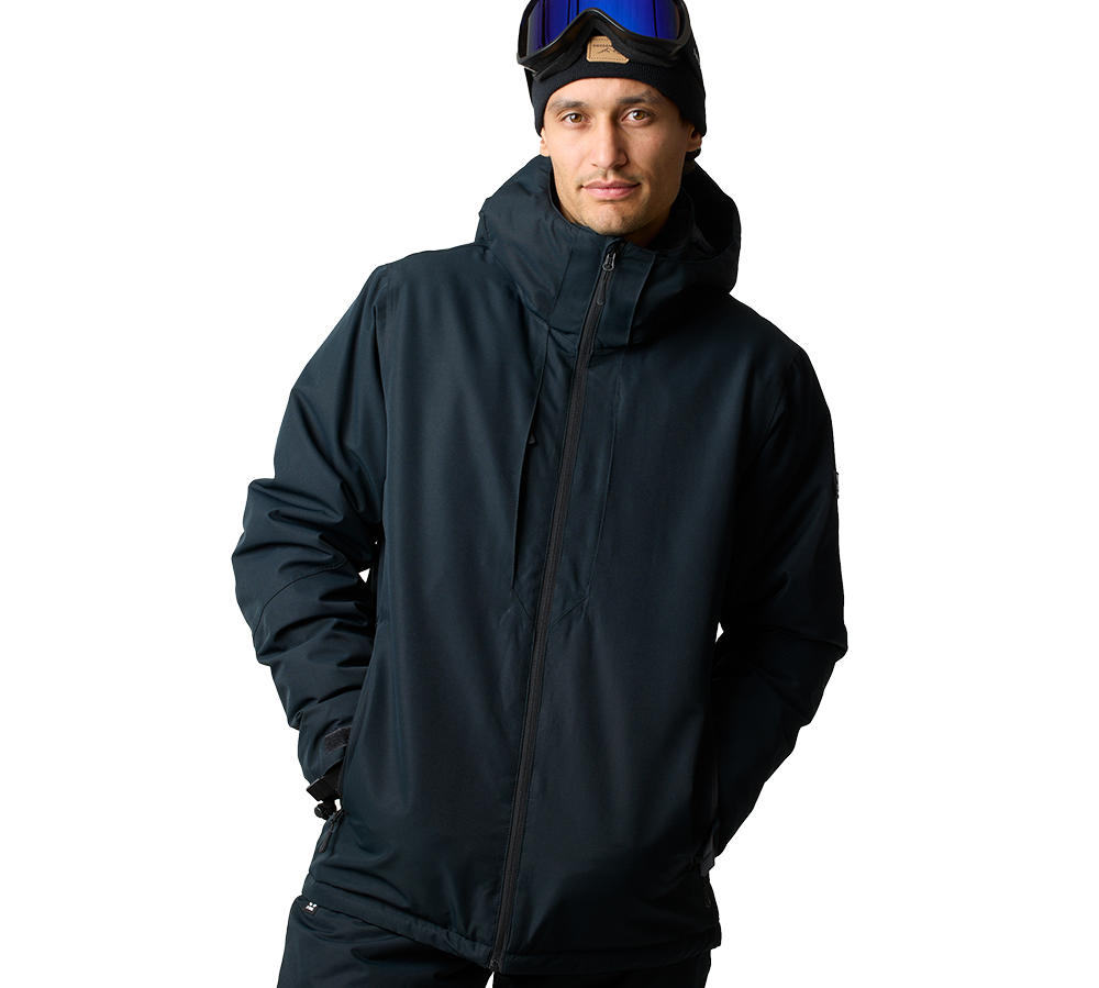 En mand iført en mørk, vandtæt skijakke med høj krave og hætte ser roligt mod kameraet. Han bærer en sort beanie og har skibriller med blåt spejlglas placeret over panden. Jakken er praktisk og isolerende med lynlås og sidelommer, og hans hænder er gemt i lommerne. Det neutrale, lyse baggrund fremhæver tøjets funktionelle og stilrene udtryk.