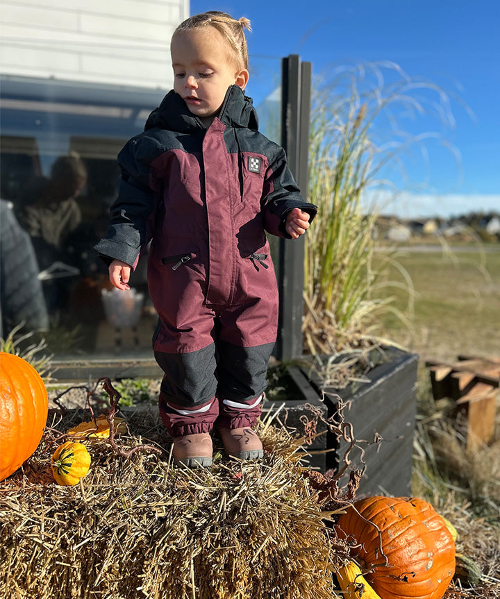 På billedet ser vi X-TRAIL Cortina Overall Jr, en flyverdragt fra X-Trail, designet til børn. Denne dragt er i mørkerøde og mørkeblå farver, og har praktiske detaljer som justerbare stropper og reflekterende elementer for øget sikkerhed. Flyverdragten ser robust og komfortabel ud, velegnet til vinterbrug og udendørs aktiviteter i koldt vejr. Den lille model står selvsikkert og demonstrerer dragtens gode pasform og fleksibilitet.