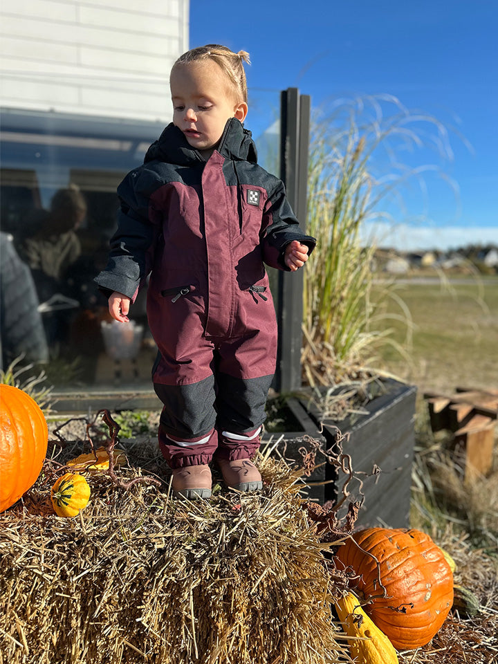På billedet ser vi X-TRAIL Cortina Overall Jr, en flyverdragt fra X-Trail, designet til børn. Denne dragt er i mørkerøde og mørkeblå farver, og har praktiske detaljer som justerbare stropper og reflekterende elementer for øget sikkerhed. Flyverdragten ser robust og komfortabel ud, velegnet til vinterbrug og udendørs aktiviteter i koldt vejr. Den lille model står selvsikkert og demonstrerer dragtens gode pasform og fleksibilitet.